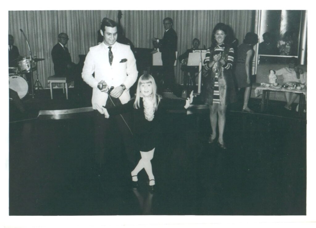 The author, aged five, onstage and curtseying after singing "Edelweiss" from the movie, "The Sound of Music." The emcee stands behind the confident, smiling child. The photo is black and white. The little girl is wearing a black velvet dress with white ruffles from her neck to her ribs and white tights and black mary-jane shoes. 